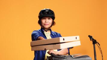 stänga upp av kurir kvinna isolerat över orange studio bakgrund föra pizza till klient. asiatisk cykel ryttare leverera hämtmat mat till kund, tar måltid lådor ut av termisk väska foto