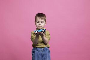 liten Lycklig barn spelar med en blå bil leksak i främre av kamera, leende litet barn njuter speltid aktivitet mot rosa bakgrund. små ung unge har roligt med färgrik fordon. foto