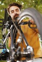 närbild av sportälskande caucasian man arbetssätt i hans gård till fixera cykel delar med specialiserade verktyg. detaljerad bild av ung manlig cyklist inspekterande och justeras cykel hjul och trampa. foto