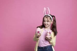 leende Lycklig barn Framställ med en rosa ägg och en kanin på kamera, bär pigtails och fluffig kanin öron i studio. glad ung flicka som visar henne påsk arrangemang och leksaker. foto