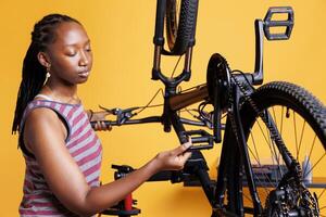 ungdomlig afrikansk amerikan kvinna försiktigt inspekterar de pedaler och redskap till göra Säker de cykel är redo för utomhus- aktiviteter. sportig afrikansk amerikan kvinna innehav och kontroll fot broms av cykel. foto