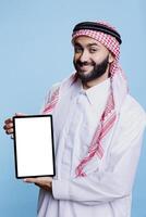 leende arab klädd i traditionell klädsel innehav digital läsplatta med vit tom skärm attrapp och ser på kamera. muslim man främja bärbar grej tömma pekskärm för Ansökan annonser foto