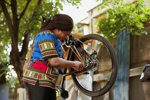 svart kvinna utomhus innehav och fixering cykel hjul med professionell utrustning. ung afrikansk amerikan kvinna reparation bruten cykel däck med arbete verktyg i gård. foto