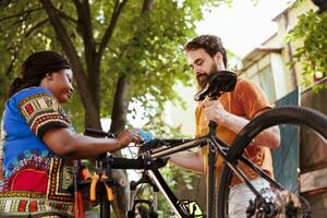 friska afrikansk amerikan lady bistå henne caucasian pojkvän fixera hans cykel utanför. aktiva ung svart kvinna fastsättning cykel till reparationsställ för atletisk man till reparera använder sig av expert- verktyg. foto