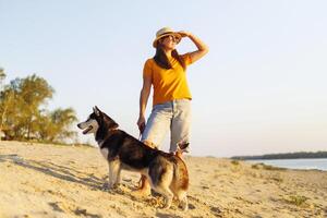 kvinna i hatt och solglasögon med henne hund njuter solnedgång på de strand foto