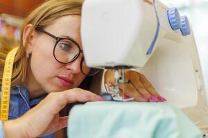 kvinna sömmerska i glasögon Arbetar på symaskin på henne arbetsplats i verkstad foto