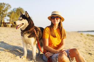 kvinna i hatt och solglasögon sitter med henne hund på de strand och njuter solnedgång foto