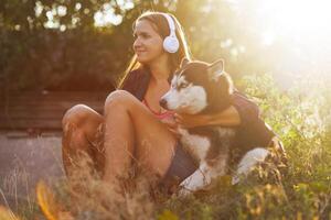 skön ung kvinna i hörlurar spelar, kramar med henne hund utomhus på solnedgång foto