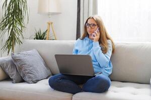 kvinna frilansare i glasögon använder sig av telefon och bärbar dator medan Sammanträde på soffa foto