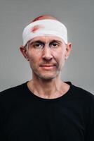 mot en grå bakgrund står en misshandlade och misshandlade man i en svart t-shirt med hans huvud insvept i en bandage foto