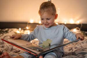 liten flicka läsning pop- upp bok i Hem säng i jul miljö med lampor bakgrund foto