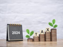 2024 skrivbord kalender, trä- blockera med ord beskatta och växter växande upp på stack av mynt. de begrepp av sparande pengar för betalning beskatta, finansiell, investering och företag växande i år 2024. foto