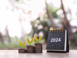 2024 skrivbord kalender, växter växande upp på stack av mynt. de begrepp av sparande pengar, finansiell, investering och företag växande i år 2024. foto