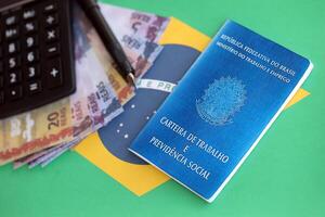 brasiliansk arbete kort och social säkerhet blå bok och reais pengar räkningar med kalkylator och penna på flagga av federativ republik av Brasilien foto