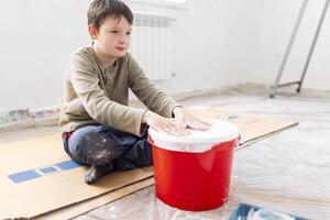 barn har roligt Sammanträde på de golv och slår hans händer på en hink av måla foto