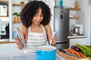 Foto av Söt afrikansk amerikan kvinna innehav matlagning slev sked medan matlagning soppa med färsk grönsaker i kök på Hem