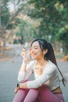 en ung kvinna åtnjuter en fredlig ögonblick, innehav en klar vatten flaska med en mild leende, som hon tar en hydratisering ha sönder under ett utomhus- träna, bär en smart klocka. foto