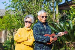 porträtt av en romantisk äldre asiatisk man spelar ukulele med hans fru medan stående i en trädgård. senior par leende och ser på de kamera. begrepp av åldrig människor och sjukvård foto