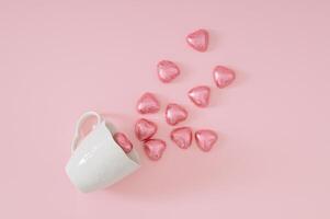 kreativ kärlek sammansättning tillverkad med rosa folie insvept choklad hjärtan kommande ut av vit kaffe kopp på pastell rosa bakgrund. minimal kärlek begrepp. romantisk choklad hjärtan aning. foto