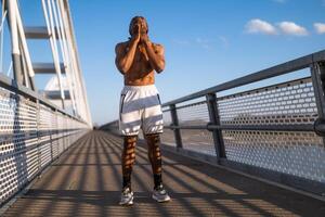 ung afrikan-amerikan man är utövar på de bro i de stad. han är stretching hans kropp. foto