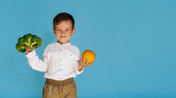 en studio skott av en pojke innehav färsk broccoli och ett orange på en blå bakgrund med en kopia av de Plats. de begrepp av friska bebis mat. foto