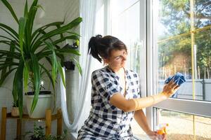 kvinna manuellt tvättar de fönster av de hus med en trasa med spray rengöringsmedel och mopp inuti de interiör med vit gardiner. återställa beställa och renlighet i de vår, rengöring serva foto