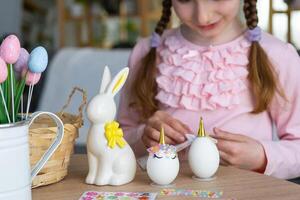 en söt flicka med rosa kanin öron gör ett påsk hantverk - dekorerar ett ägg i de form av en enhörning med strass, horn, blommor i de interiör av en hus med växter. foto