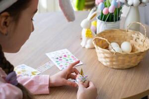 en söt flicka med rosa kanin öron gör ett påsk hantverk - dekorerar ett ägg i de form av en enhörning med strass, horn, blommor i de interiör av en hus med växter. foto