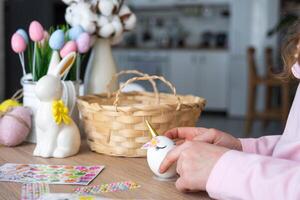 en söt flicka med rosa kanin öron gör ett påsk hantverk - dekorerar ett ägg i de form av en enhörning med strass, horn, blommor i de interiör av en hus med växter. foto