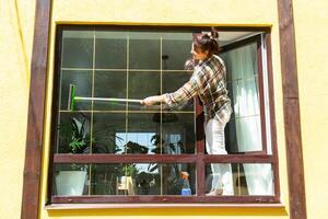en kvinna manuellt tvättar de fönster av de hus med en trasa med en spray rengöringsmedel och en mopp utanför. säkerhet på höjd, återställa beställa och renlighet i de vår, rengöring service foto