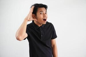 ung asiatisk man ser kamera och håller på med kan inte höra gest, påfrestande till lyssnande en skvaller bär svart polo t skjorta isolerat på vit bakgrund foto