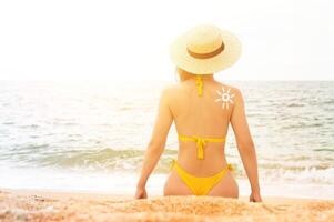 en ung caucasian kvinna i en baddräkt och en sugrör hatt sitter på de strand i främre av de hav med henne tillbaka i de ram. de Sol på en kvinnas tillbaka är dragen med Solskydd foto