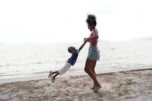 blandad lopp afrikansk och asiatisk mor och pojke är spelar på de utomhus- område. leende Lycklig familj ha roligt löpning på de strand. porträtt av mamma och unge livsstil med en unik frisyr. foto