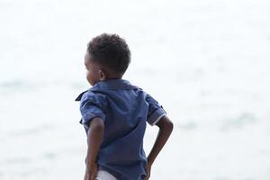 blandad lopp afrikansk och asiatisk pojke är spelar på de utomhus- område. leende Lycklig pojke har roligt löpning på de strand. porträtt av pojke livsstil med en unik frisyr. foto