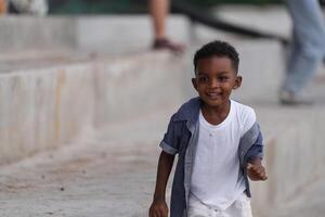 blandad lopp afrikansk och asiatisk pojke är spelar på de utomhus- område. leende Lycklig pojke har roligt löpning på de strand. porträtt av pojke livsstil med en unik frisyr. foto