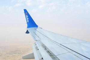 se av en blå flygplan vinge med de logotyp flydubai mot en dimmig öken- bakgrund från ett flygplan fönster. dubai, uae - augusti 15, 2023 foto