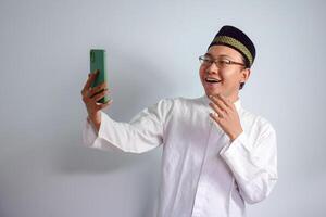 asiatisk muslim man bär glasögon och vit trasa pekande telefon utgör för Ramadhan och eid al fitr. isolerat vit bakgrund foto