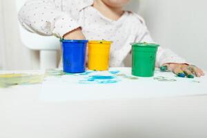 skön liten flicka drar med finger målarfärger på en vit ark av papper. kreativ barn utveckling i dagis eller fri tid på Hem foto