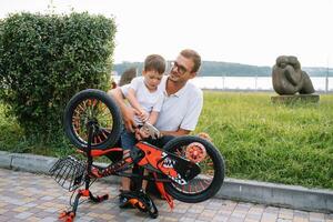 Lycklig far och hans son har roligt tillsammans på de grön parkera, fixering cykel tillsammans. fars dag. foto