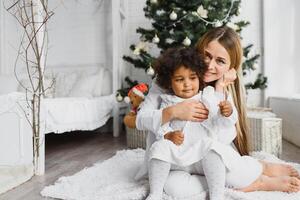 glad jul och Lycklig högtider glad mamma och henne söt dotter på jul träd. förälder och liten barn har roligt nära jul träd inomhus. kärleksfull familj med presenterar i rum. foto