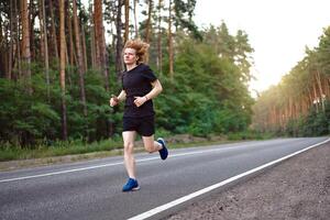 caucasian ung lockigt man idrottare kör solig sommar dag på asfalt väg i de skog. foto