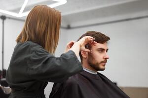 stilig blå ögon man Sammanträde i barberare affär. hårstylist frisör kvinna skärande hans hår. kvinna barberare. foto