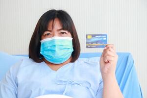 fet asiatisk kvinna patient bär en mask i en sjukhus patient rum. hon innehar en kreditera kort till betala. medicinsk service begrepp. patienter smittad med coronavirus. hälsa försäkring. foto