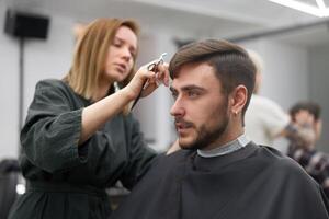 stilig blå ögon man Sammanträde i barberare affär. hårstylist frisör kvinna skärande hans hår. kvinna barberare. foto