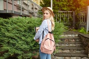 ung studerande blond i en sugrör hatt och en rosa ryggsäck är stående på de främre av de universitet och innehav bärbara datorer foto