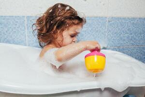 skön liten flicka tar en bad på Hem. en söt bebis är Sammanträde i de badrum och spelar med leksaker och vatten. personlig hygien för barn. dagligen hygien foto
