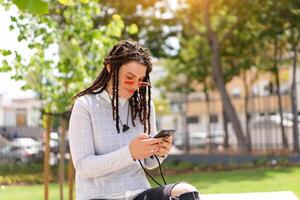 ung kvinna hipster med dreadlocks i rosa solglasögon använda sig av smartphone Sammanträde sommar parkera utomhus. ny generation, teknologi i hand. foto