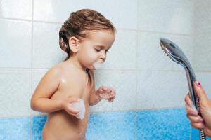 skön liten flicka tar en bad. en barn är spelade med vatten och spray från de dusch. glad barns hygien. stygg dotter i de badrum foto