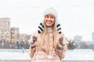 Lycklig vinter- tid i stor stad charmig flicka stående gata klädd rolig fluffig hatt. njuter snöfall, uttrycker positivitet, leende till kamera foto