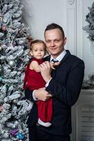 pappa i en företag kostym innehar hans dotter klädd i ett elegant röd klänning på de bakgrund av de jul träd foto